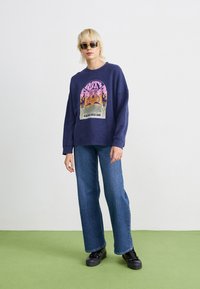 Sweat-shirt marine avec un imprimé graphique de montagnes et de flammes, associé à un jean bleu à jambes larges et à des baskets noires à plateforme. Les accessoires comprennent des lunettes de soleil.