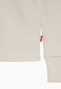Langermet topp i vaffelstrikket stoff i lyse beige med ribbede mansjetter. Har en rød Levi's-lapp sydd på siden. Mykt, teksturert stoff.