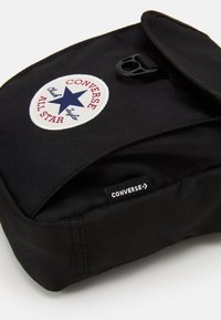 Mochila de tecido preto com um emblema redondo branco contendo uma estrela azul e texto vermelho. Inclui uma aba e um rótulo da marca na lateral.