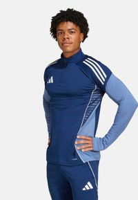 Marineblauw sportshirt met halve rits, lange mouwen en drie witte strepen op de schouders. Bevat lichtblauwe zijkanten met textuur.