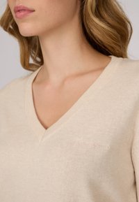 Pull en maille beige avec un col en V, présentant une texture délicate et un tissu doux. Comprend un subtil logo brodé en rose pâle sur la poitrine.