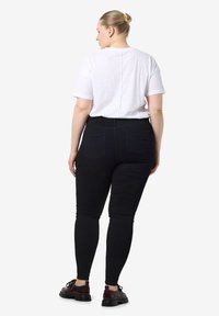 Mujer con cabello rubio recogido en un moño, vistiendo una camiseta blanca de manga corta, jeans ajustados negros y zapatos marrón oscuro, de pie de espaldas.