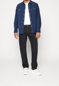 Camisa azul marino de botones con dos bolsillos delanteros, usada sobre una camiseta blanca, combinada con pantalones negros y zapatillas blancas.