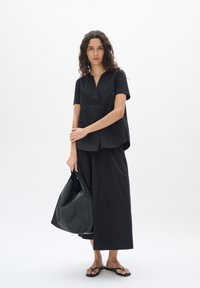 Top nero a maniche corte con scollatura a V e vestibilità ampia, abbinato a pantaloni larghi coordinati. Una borsa in pelle nera è portata sotto il braccio.