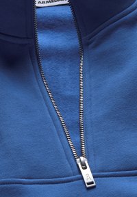 Pull-over en polaire bleu avec demi-fermeture éclair, doté d'une fermeture éclair en métal et d'une texture lisse, avec une bande bleu légèrement plus foncée au niveau du col.