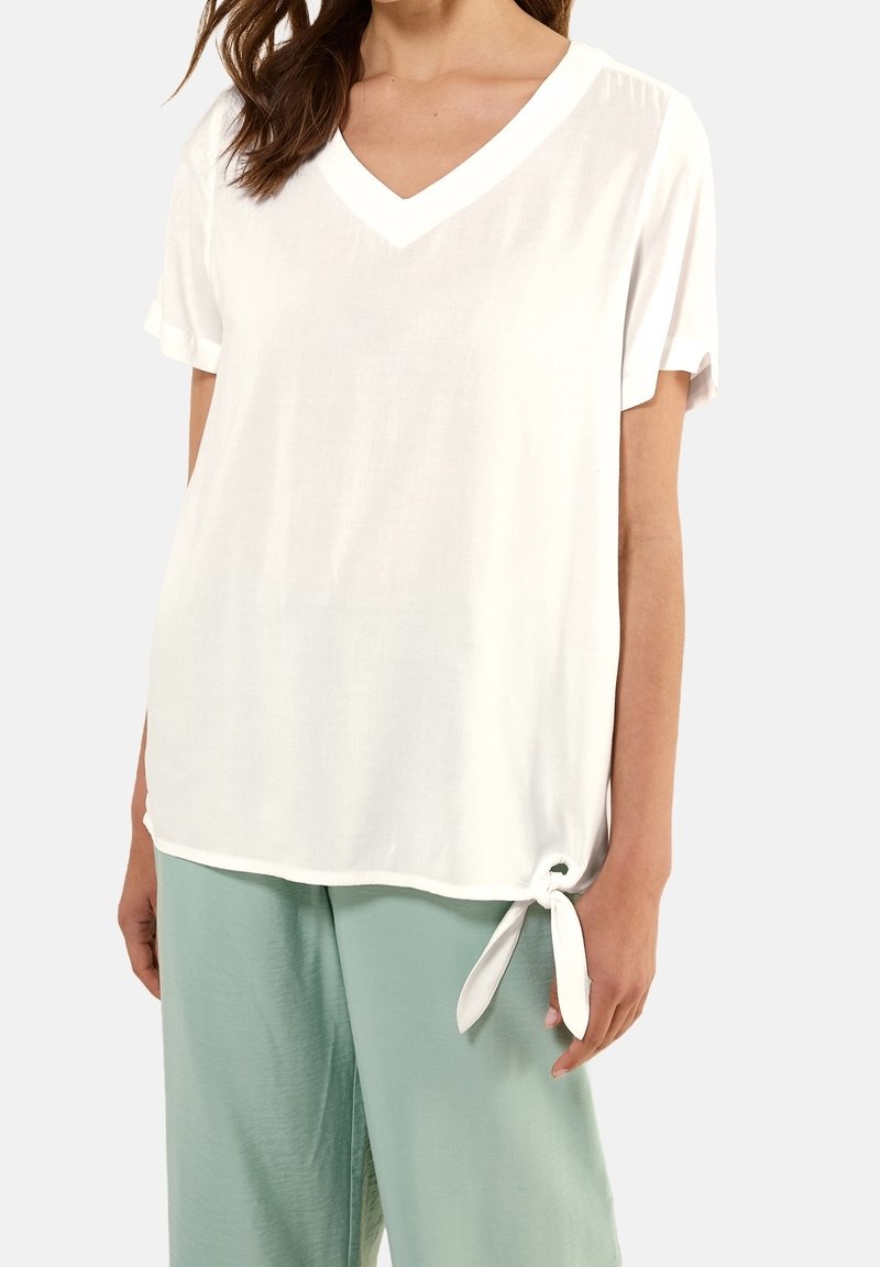 Femme portant un haut blanc ample à manches courtes avec un col en V et un détail de nouage sur le côté, associé à un pantalon large vert clair.