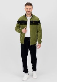 Chaqueta de terciopelo verde con franjas negras y logo dorado, combinada con pantalones de chándal negros con franjas laterales blancas, llevada con zapatillas blancas.