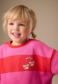 Sudadera con rayas horizontales rojas y rosas, cuello redondo y diseño bordado de cerezas con el texto "BEARY SWEET". Textura de tela suave.