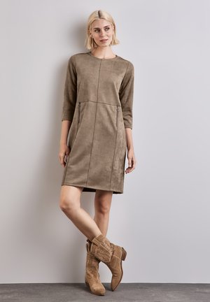 Robe en daim taupe à manches trois-quarts, dotée de coutures contrastantes et de poches zippées. Associée à des bottes de cowboy beiges.
