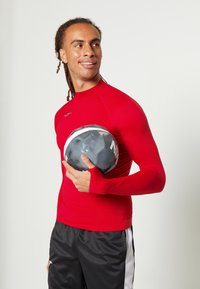 Rotes Sportshirt mit langen Ärmeln und Stehkragen, kombiniert mit schwarzen Shorts mit seitlichen Streifen. Das Modell hält einen schwarz-weißen Fußball.
