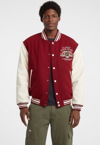 Punane ja kreemikat värvi varsity jakk ribadega mansettide ja vöökoha, millel on tikitud logo rinnal ja nööpkinnitusega ees.