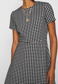 Svart- och vitrutigt mönstrat klänning med korta ärmar, figurnära överdel och utsvängd kjol. Guld-färgad lagerhalsband ger extra detalj.