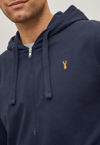 Mornarska zip-up dukserica napravljena od pamuka, s kenguru džepom i malim izvezenim dizajnom u zlatnoj boji na lijevoj strani prsnog područja.