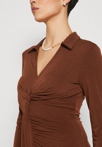 Robe marron avec un décolleté en V et un détail torsadé au niveau de la poitrine, dotée de manches longues et d'un côté froncé. Collier en perles et en chaîne dorée en accent.
