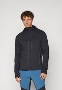 Giacca nera con cappuccio, frontale con zip, tessuto testurizzato e due tasche con zip. Indossata con pantaloni sportivi blu caratterizzati da blocchi di colore.