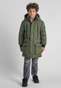 Parka acolchada de color verde oliva con capucha, cierre frontal con cremallera, dos bolsillos en el pecho y dos bolsillos inferiores con solapa, combinada con unos jeans grises desgastados y zapatos negros.