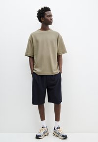 Oliven grøn oversized t-shirt, marineblå stribede shorts, hvide sokker og beige sneakers. Model står imod en ensfarvet hvid baggrund.