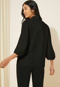 Blusa nera con colletto e maniche a sbuffo. Tessuto morbido, vestibilità rilassata e dettaglio con spacco sul retro. Abbinata a pantaloni neri attillati.