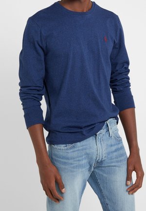Homme portant un t-shirt bleu à manches longues et col rond avec un logo rouge sur la poitrine et un jean bleu clair, debout les mains détendues sur les côtés.