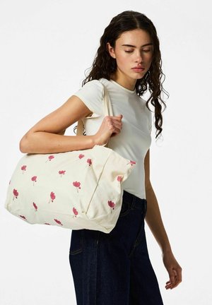 Mlada žena u bijeloj majici i tamnim trapericama nosi kremastu torbu s ružičastim cvjetnim uzorcima preko ramena.