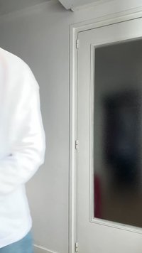 Άτομο με λευκό πουκάμισο, μερικώς ορατό, περπατά δίπλα από κλειστή λευκή πόρτα με γαλακτερό γυαλί σε δωμάτιο με γκρι τοίχους.