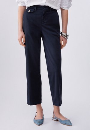 Persona che indossa pantaloni dritti blu scuro, blusa bianca testurizzata, scarpe slingback blu e bracciali d’argento, in piedi contro uno sfondo semplice.
