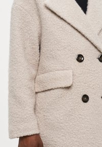 Μπεζ παλτό από μειγμα μαλλιού με υφή, αφράτη επιφάνεια, διπλή κουμπί, μεγάλες μαύρες κουμπότρυπες και μία μόνο τσέπη μπροστά.