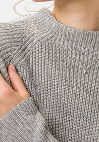 Pull gris en maille avec texture côtelée et larges poignets, doté d'un col rond. Un délicat collier argenté est visible, tombant doucement.