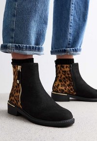 Černé semišové kotníkové boty s leopardím potiskem na zadní části, zlatými zipovými detaily, nošené s ohrnutými modrými džíny a černými ponožkami na světlé podlaze.