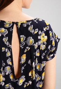 Blouse florale marine à manches courtes, ornée de fleurs jaunes et grises, avec un dos plissé et une ouverture en forme de clé sécurisée par un bouton noir.