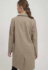 Cappotto in lana beige con colletto, maniche lunghe e taglio a lunghezza media. Presenta una cucitura centrale sul retro e una texture liscia.