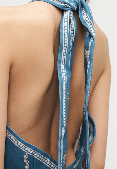 Μπλε κορυφή με σχέδια, δεμένη στο λαιμό, που αφήνει ακάλυπτα τα πίσω και τον ώμο με ανοιχτόχρωμο δέρμα, πάνω σε απλό φόντο.