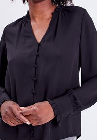 Blusa negra con escote en V, mangas abullonadas y puños con volantes. Presenta un cierre de botones en la parte delantera con botones forrados de tela y una textura suave.