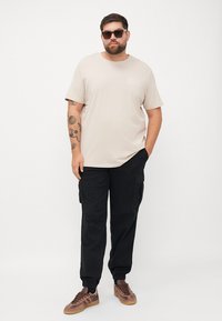 Beige katoenen t-shirt met een klein logo op de borst, gecombineerd met zwarte cargo pants met zijzakken en elastische boorden, en bruine sneakers.