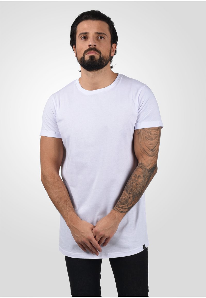 Weißes Baumwoll-T-Shirt mit kurzen Ärmeln, Rundhalsausschnitt und längerem Schnitt. Verfügt über ein kleines schwarzes Etikett am Saum und eine glatte Textur.