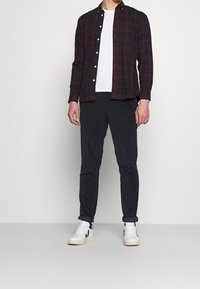 Červeno-černá kostkovaná košile na knoflíky přes bílém triku, doplněná tmavě šedými kalhotami a bílými teniskami s černými akcenty.