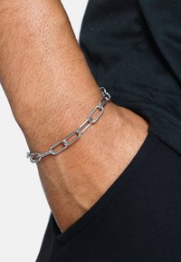 Pulsera de cadena de plata con eslabones alargados, llevada en una muñeca. Presenta metal pulido y un acabado brillante, que contrasta con la tela más oscura.