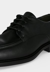 Gros plan d'une chaussure habillée en cuir noir avec un bout arrondi, un laçage à l'avant et un talon bas empilé sur fond blanc.