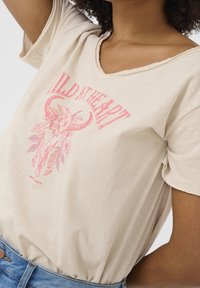 Béžové tričko s krátkým rukávem a výstřihem do V, s růžovým grafickým designem býčí lebky obklopené květinami a textem "Wild N Heart."