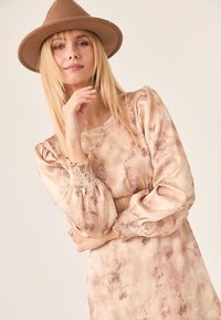 Vestido de manga larga, beige, con un patrón de tie-dye, puños fruncidos y escote redondo. Combinado con un sombrero marrón de ala ancha.