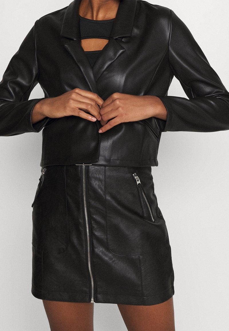 Musta keinonahkainen lyhyt takki lovitetulla kauluksella, yhdistettynä mustaan keinonahkaiseen minihameeseen, jossa on vetoketjulliset taskut ja etukiinnitys.