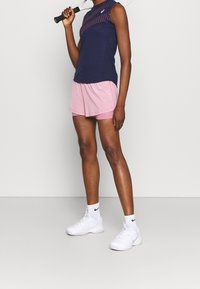 Haut de sport sans manches bleu marine avec des accents roses, associé à un short rose. Le modèle porte des chaussures de sport blanches et des chaussettes de cheville.