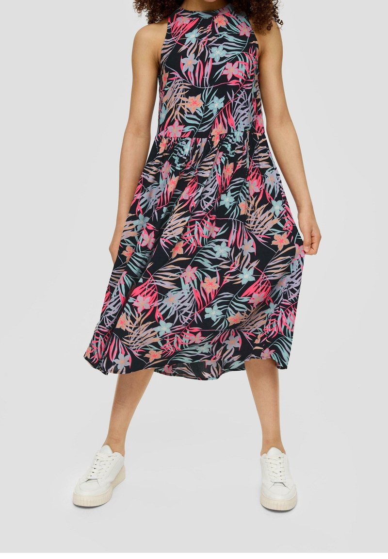 Robe midi à col licou en tissu noir avec un imprimé coloré de feuilles tropicales et de fleurs. Portée avec des baskets blanches, montrant une coupe décontractée.