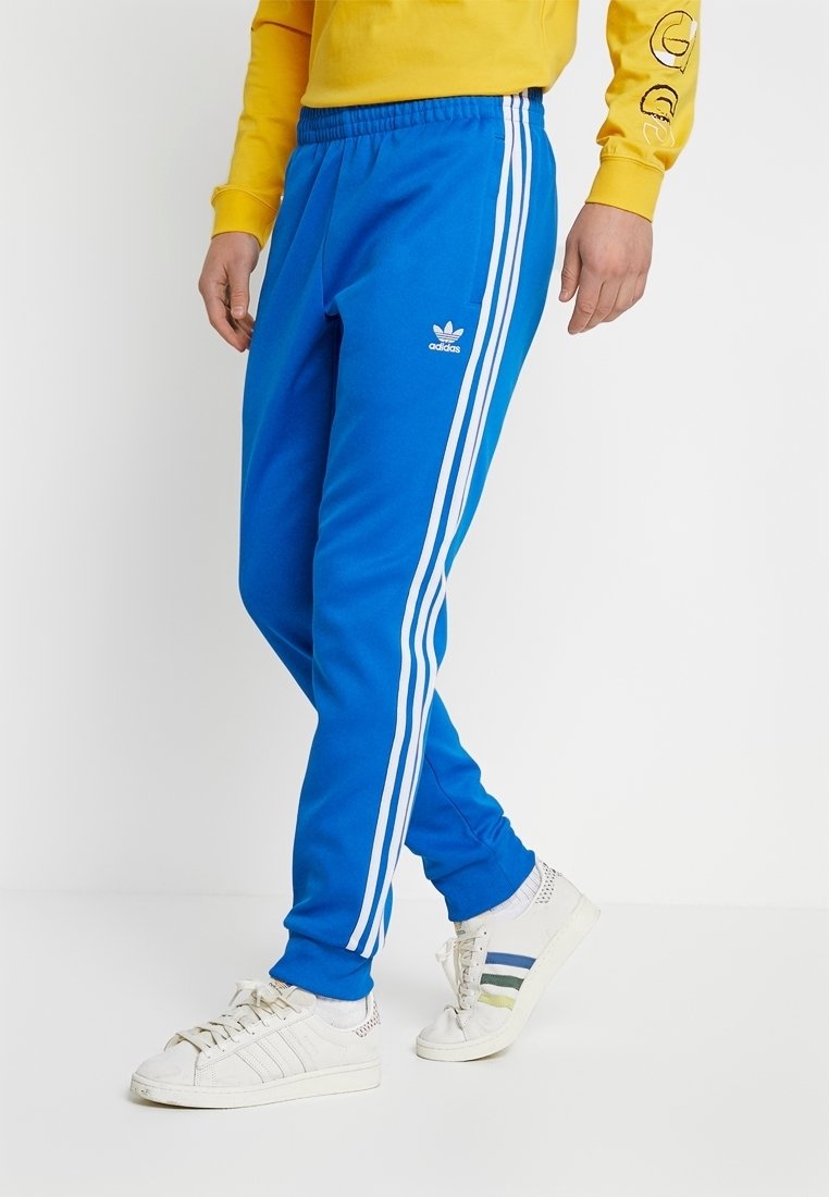 Blå adidas trackbyxor med vita sidostripor, elastisk midja och mjukt tyg. Matchas med vita sneakers med färgade ränder.
