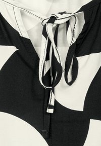 Haut en tissu noir et blanc avec un décolleté en V, fermeture nouée avec un cordon de serrage, et de grands motifs géométriques. Détail de surface texturée visible.