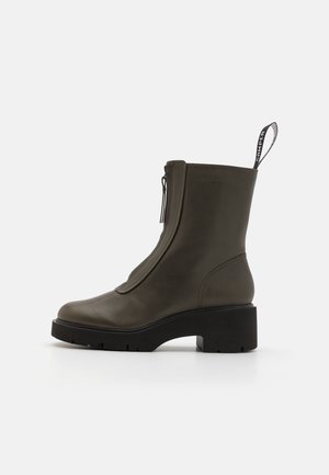 Olivově zelená kožená kotníková bota s hladkým povrchem, bočním zipem a silnou černou podrážkou. Obsahuje poutko na zadní straně.