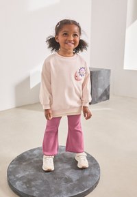 Ljusrosa sweatshirt med puffärmar, prydd med broderier av sol och måne; kombinerad med rosa utsvängda byxor och vita sneakers på en mörk rund piedestal.