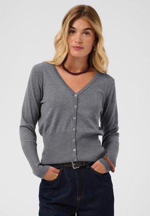 Femme aux cheveux blonds portant un cardigan gris boutonné, un jean foncé, un bracelet marron et un collier de perles rouge foncé, les mains dans les poches, fond neutre.
