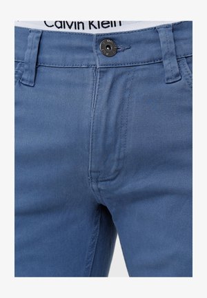 Jeans in denim azzurro chiaro con vestibilità classica, chiusura con bottone in metallo e vita bianca con logo del brand nero. Texture liscia e cuciture pulite.