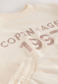 Krém színű pulóver bordázott nyakkal, puha, domború hímzéssel, amely a "COPENHAGEN 1997" feliratot világos bézs színben mutatja. Látható gumis derékrész részlet.